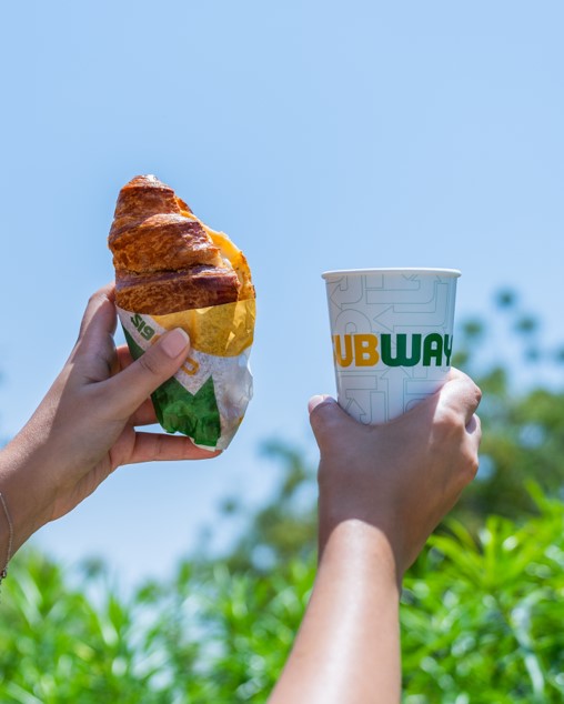Hands holding a Subway croissant sandwich and drink cup against a blue sky